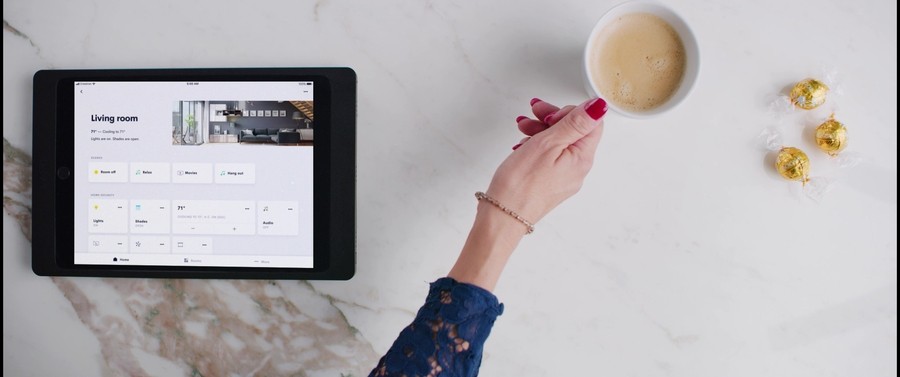 Hand reaching for a cup of coffee on a counter with a tablet displaying a smart home interface. 